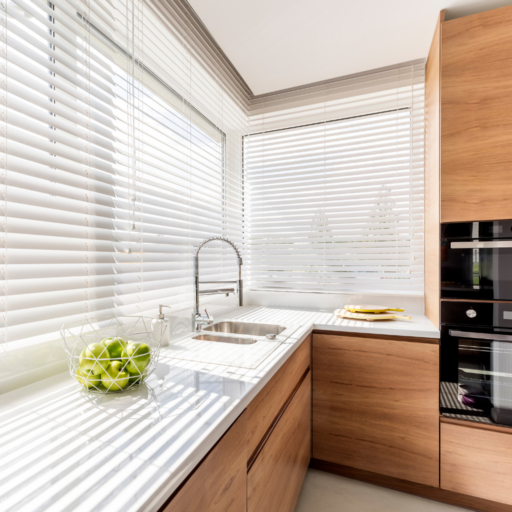 a view to a sunny kitchen, kitchen is made of wood with white top panels. Windows have white aluminum blinds. There is a basket with green apples  near the sink. 