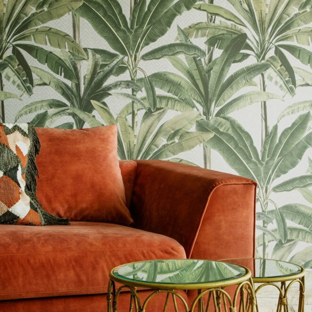 big green plam leaves on a wallpaper, an orange velvet couch in front and two bamboo tables.
